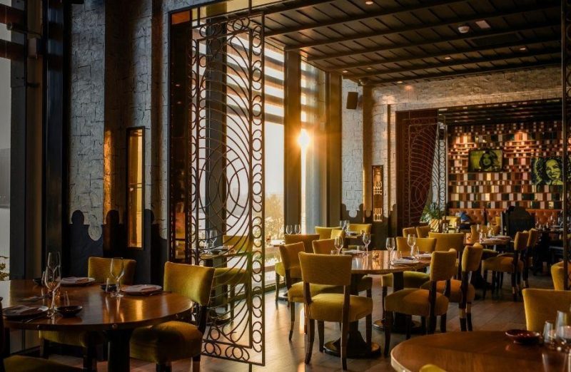 Sophisticated restaurant interior bathed in golden sunset light. It features mustard yellow velvet chairs, round wooden tables, and an intricate laser-cut metal screen. The walls are made of textured grey stone with colorful portraits.