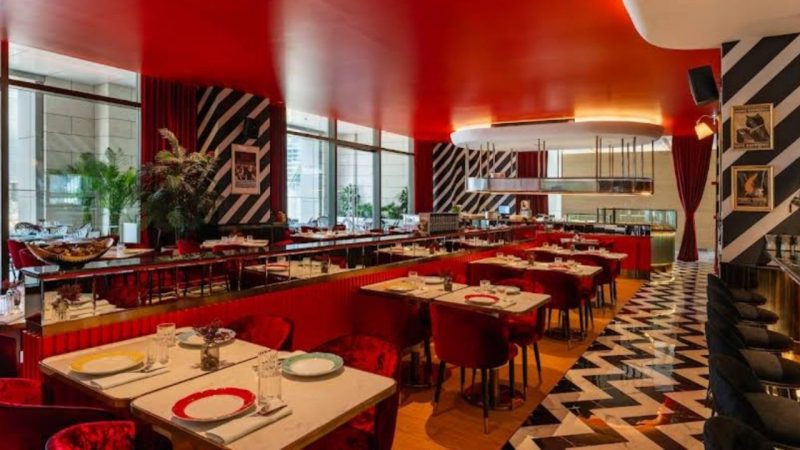 High-energy retro-chic restaurant interior with a bold red ceiling and black-and-white zigzag patterned flooring. The space is filled with red velvet seating, mirrored partitions, and striped pillars.