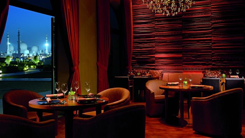 Dramatic fine-dining interior at night with deep red velvet curtains and a textured red feature wall. An open doorway offers a stunning view of the illuminated Sheikh Zayed Grand Mosque under a twilight sky.