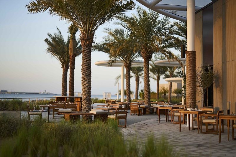 Coastal outdoor dining area with wooden furniture arranged under tall palm trees. The palm trunks are wrapped in rope, and the background shows a calm sea view and modern white architectural shade structures.