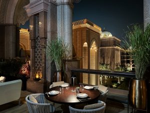 A stylish round dining table on a luxury terrace with illuminated arches and Arabian-style architecture, overlooking palm trees and domed buildings at night, creating an elegant and romantic dining atmosphere.