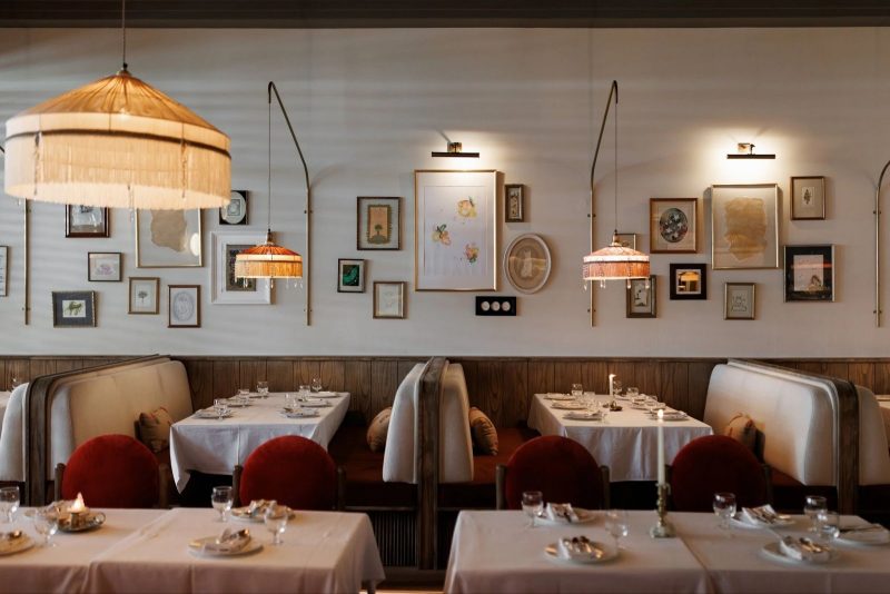 Cozy, vintage-inspired restaurant interior with a gallery wall of small framed art. White tablecloths, red velvet chairs, and wooden booths are lit by ornate tasseled fringe pendant lamps.