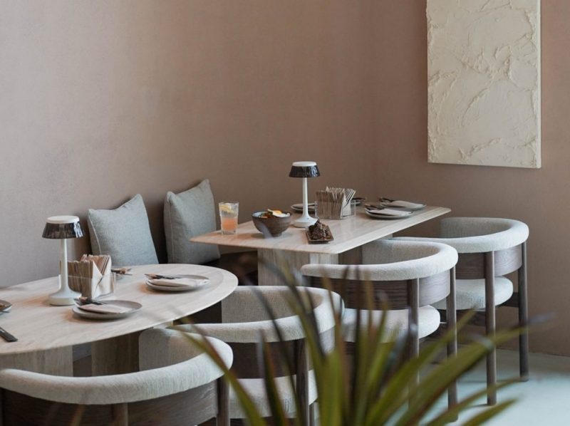 Minimalist dining area with a muted clay-colored wall. It features curved light-gray fabric chairs, marble tables, and sleek white modern table lamps. A white textured abstract artwork hangs on the wall, creating a serene atmosphere.