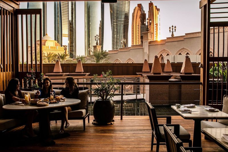 An outdoor restaurant terrace with a view of modern skyscrapers. People are dining at a marble table with dim sum baskets. The space features warm wood flooring, wooden slats, and decorative stone pillars.