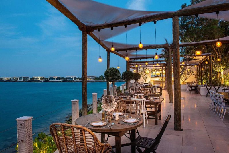 An outdoor waterfront restaurant at twilight with wooden tables, wicker chairs, and hanging Edison bulbs under a white canopy.