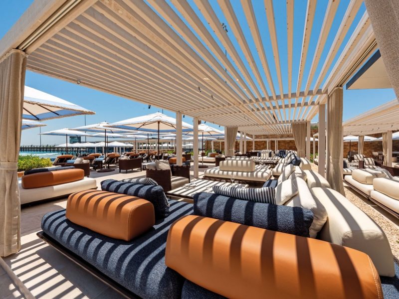Luxury outdoor beach club lounge under a wooden slat pergola. The seating area includes comfortable sofas with navy-white striped cushions and tan leather bolsters. White beach umbrellas and a clear blue sky are visible in the background.